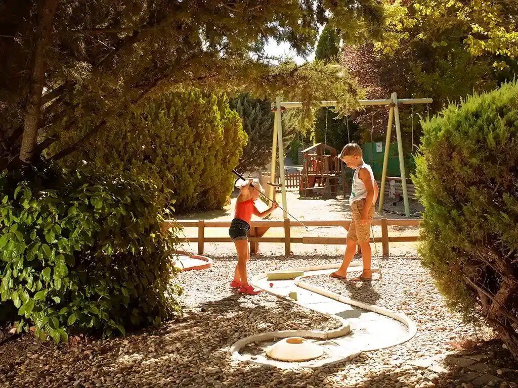 Dos personas jugando un juego en un camino de grava con una pista circular, rodeado de follaje y una valla de madera.