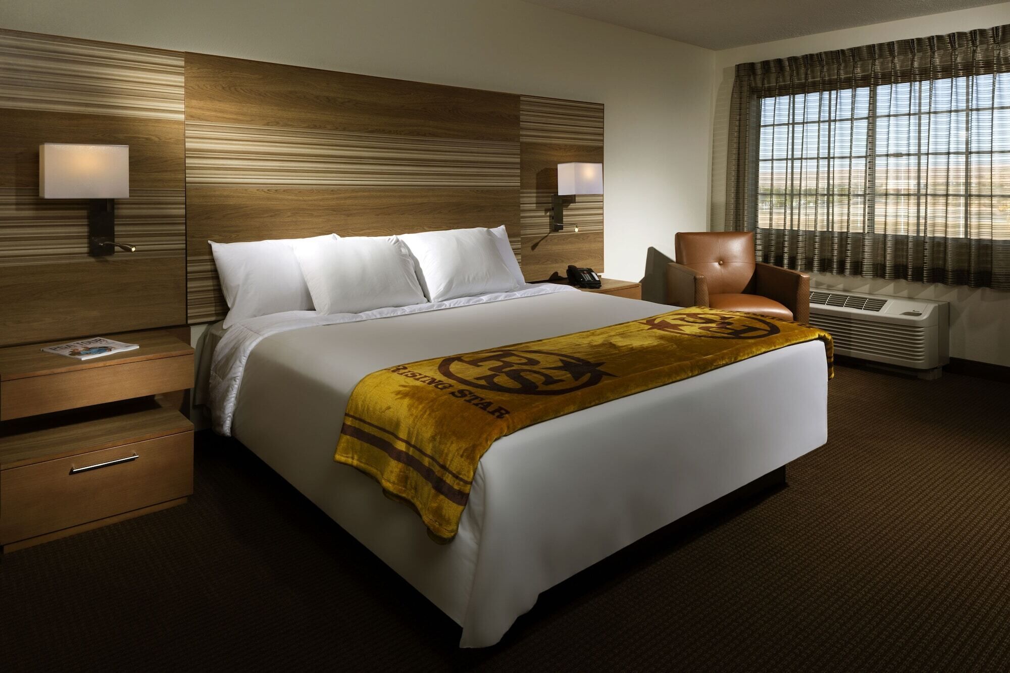 A hotel room with a large bed, two bedside tables, a chair, a window with curtains, and a wall-mounted air conditioner.
