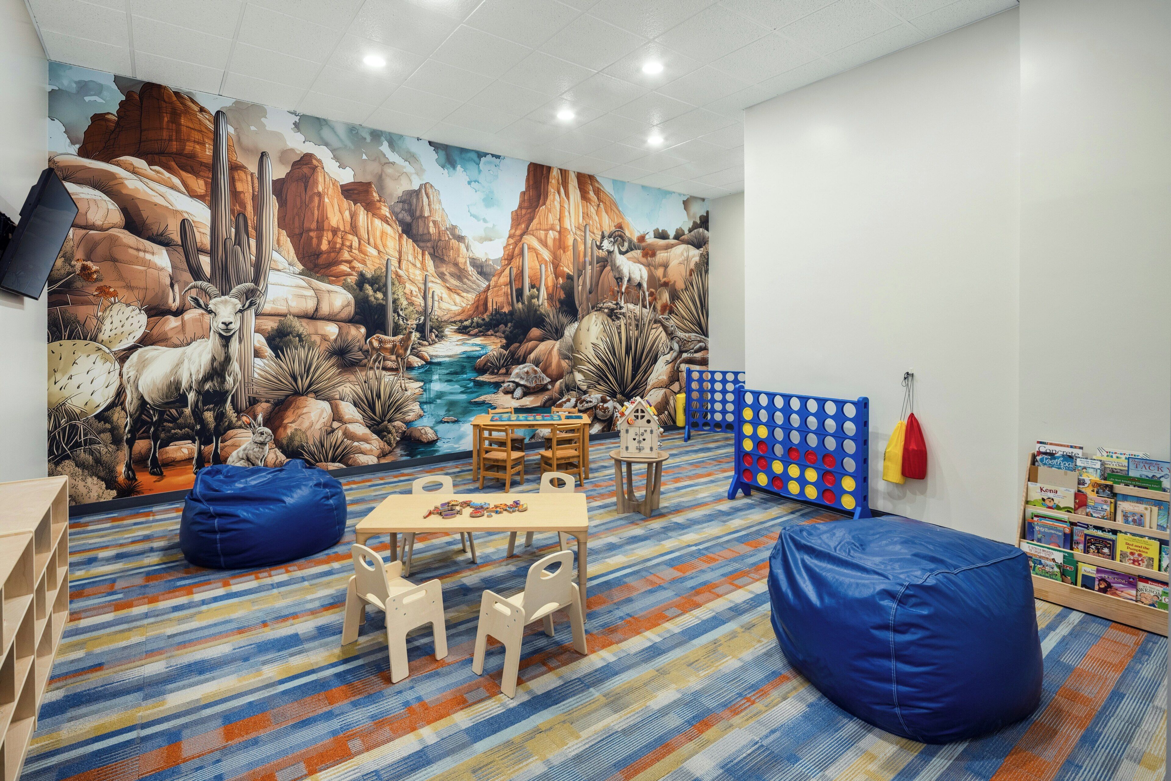 Une salle de jeux pour enfants avec une fresque murale, des tables pour jouer, des fauteuils en forme de sac et une bibliothèque.