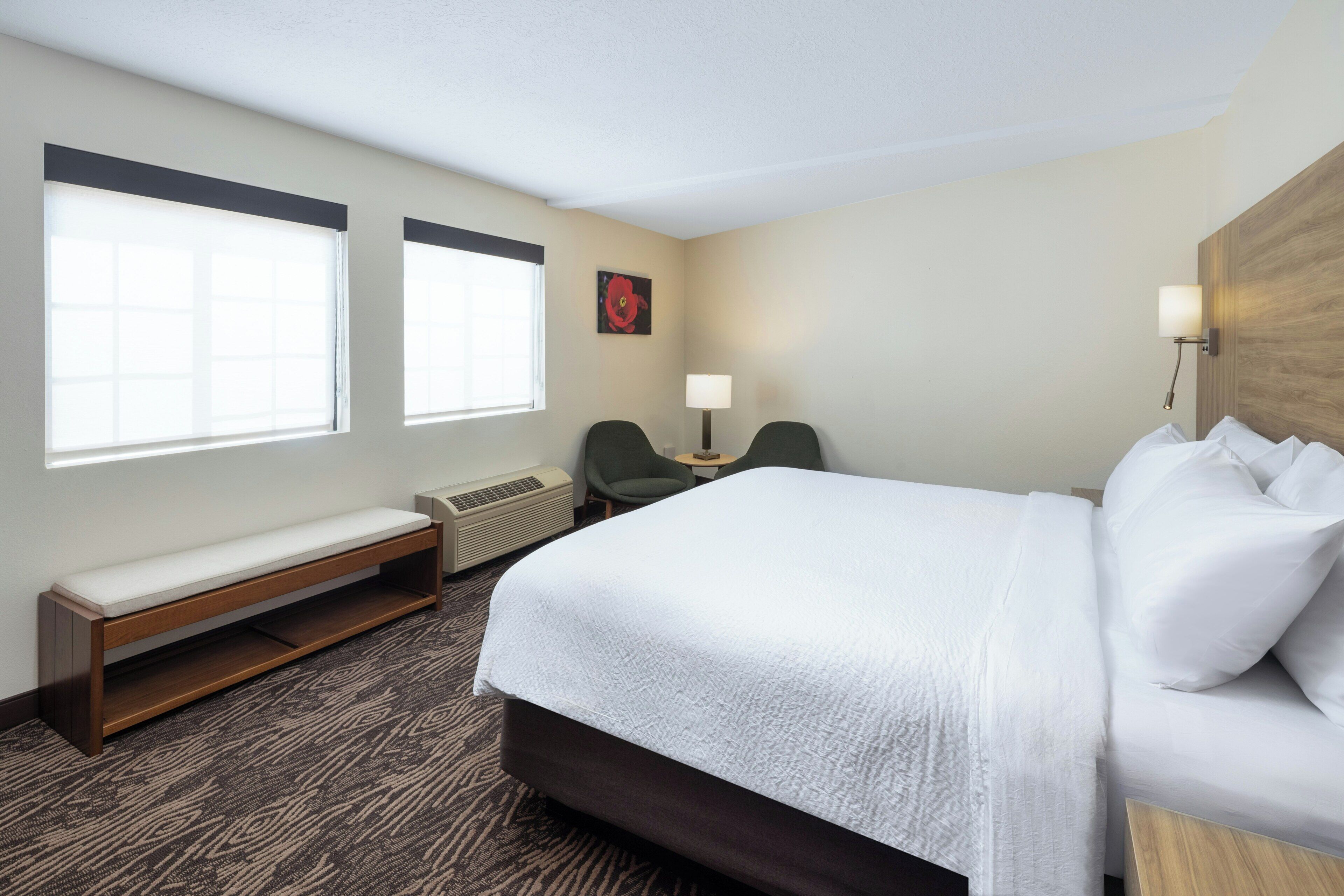 Una habitación de hotel con una cama grande, un banco, dos sillas, una mesita y una lámpara de pared.