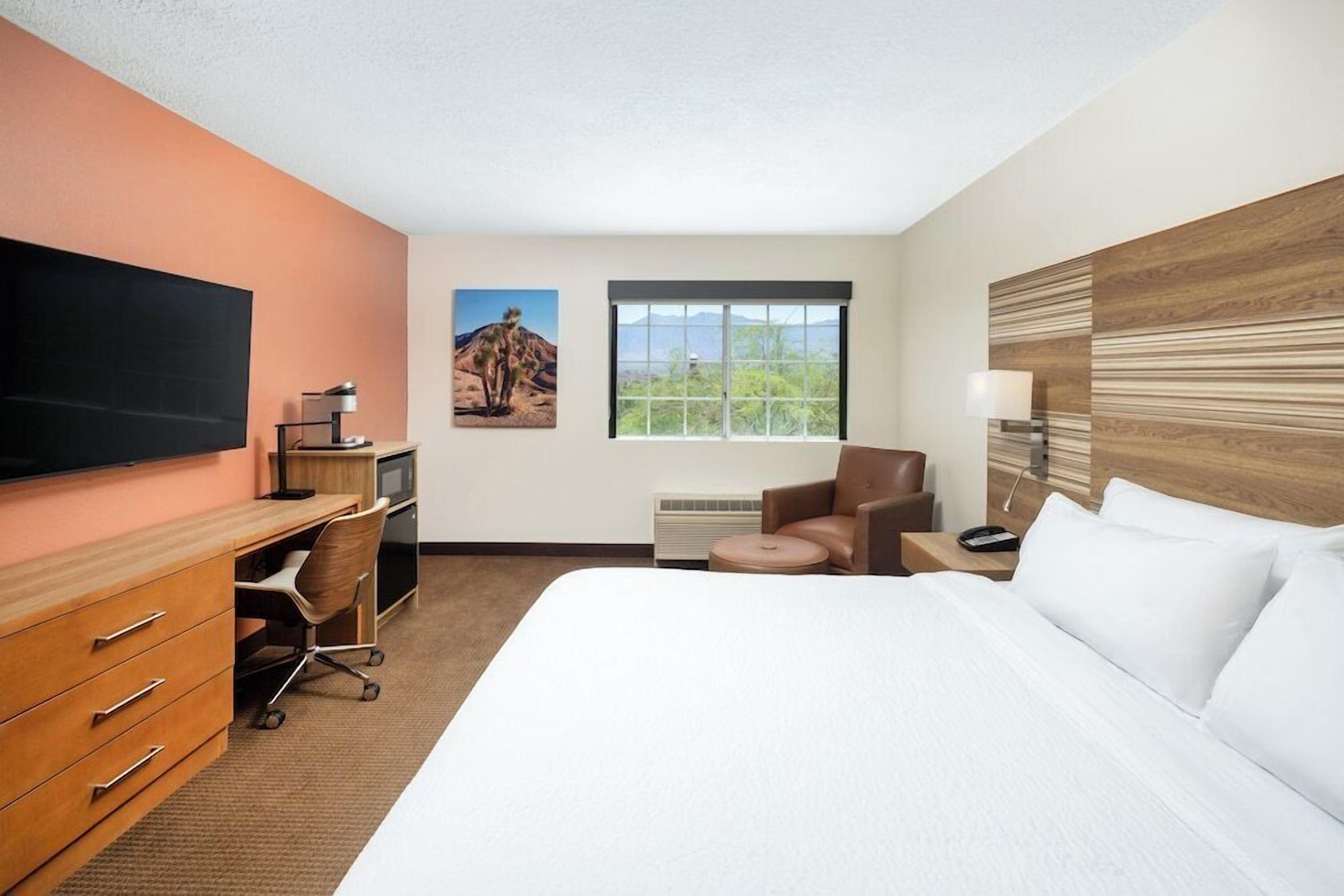 A hotel room with a large bed, a desk with a computer, a chair, a television, and a window with a view of greenery.