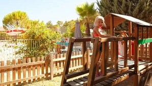 Un niño jugando en un parque infantil de madera con un tobogán y una zona de escalada, rodeado de una valla y árboles.