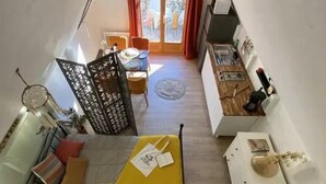 Et loft-studie med højt til loftet, spiseplads med bord og stole, tekøkken med vask og komfur, og en gul seng med puder og en lille plante.