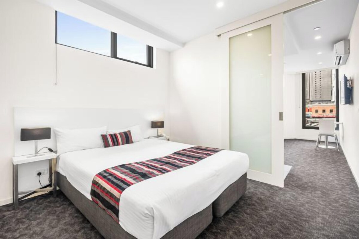 A modern hotel room with a large bed, bedside tables, a desk, and a view of the city.