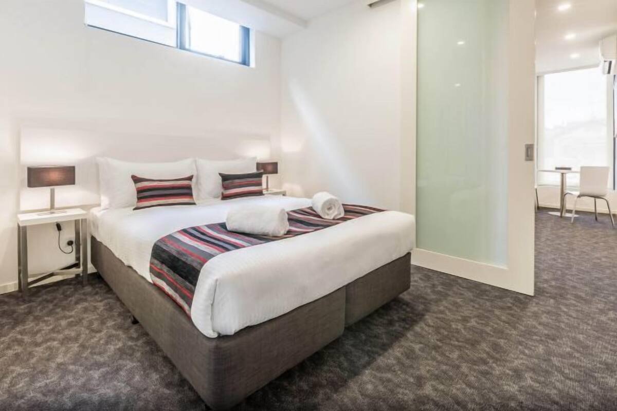 A modern hotel room with a large bed, a bedside table, a lamp, and a view of the city through a large window.