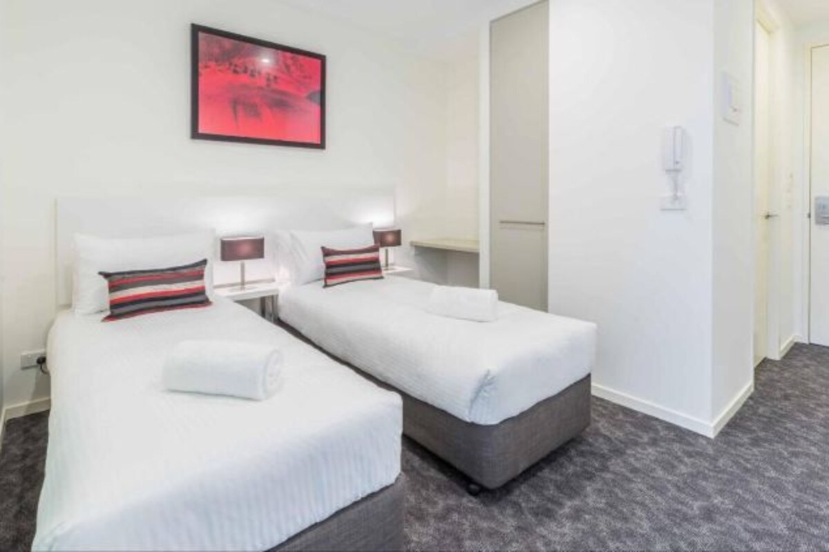 Hotellihuone, jossa on kaksi erillistä sänkyä, punainen abstrakti maalaus, valkoinen ovi ja harmaa matto.