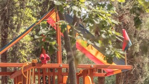 Un parque infantil con un tobogán amarillo, un columpio de cuerda y una estructura de madera para escalar.