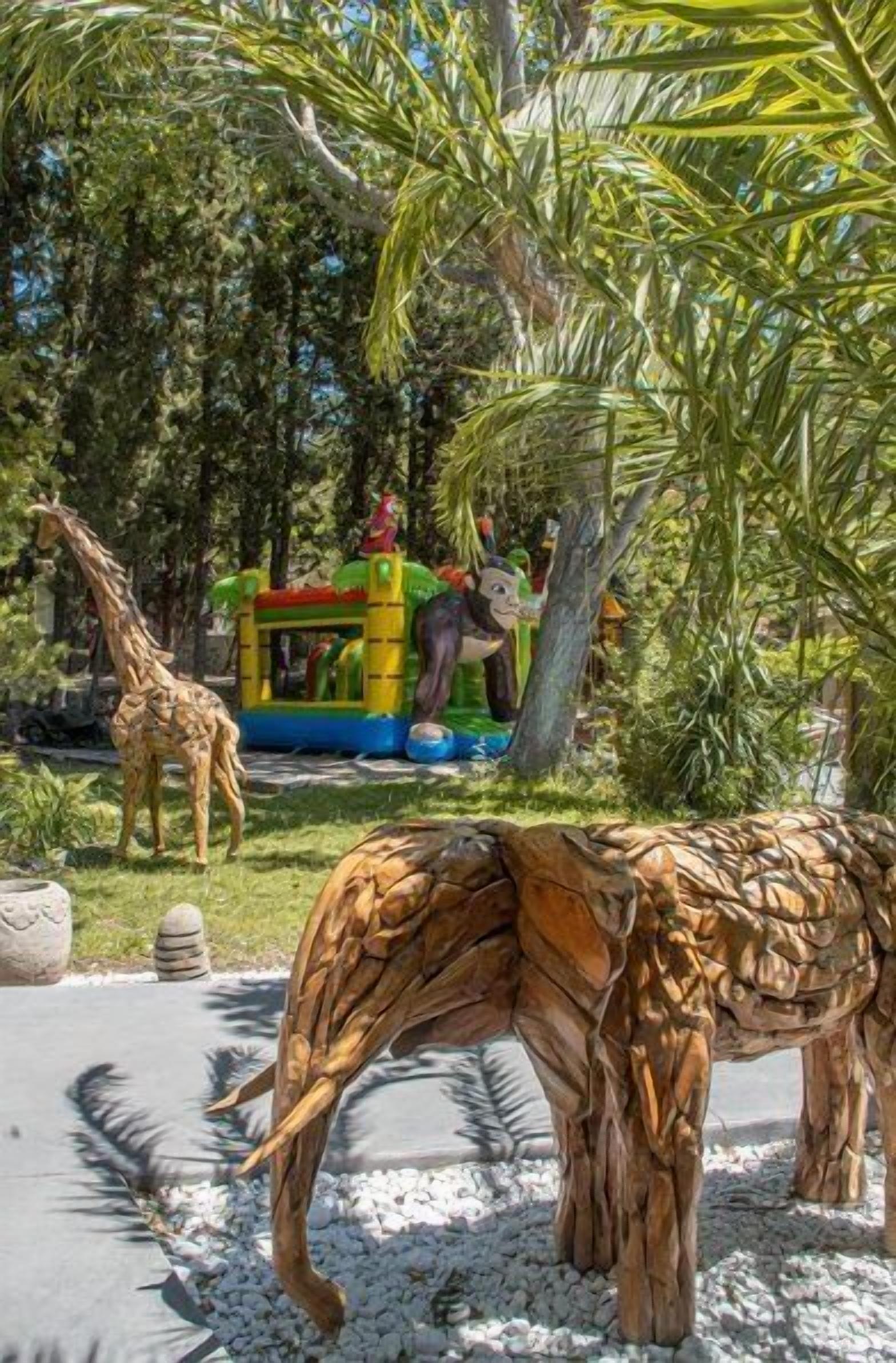 Un jardí amb una estàtua de girafa, una escultura de fusta d'elefant i una estructura de joc colorida.