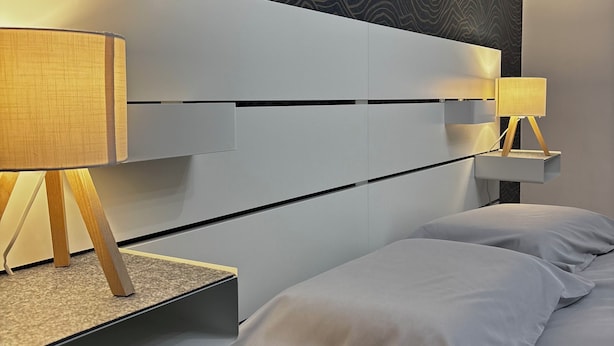 Ein modernes Schlafzimmer mit einem weißen Kopfteil, zwei Nachttischen mit Lampen und einer strukturierten Wand hinter dem Bett.