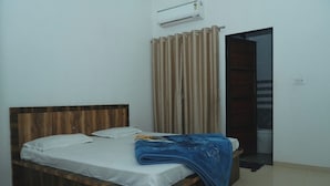 A single bed with a wooden headboard, white bedding, and a blue blanket. There is a beige curtain partially drawn, and an air conditioning unit mounted on the wall. - Hotel Vidya Dham Vrindavan  (Mathura)