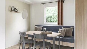 A room with a dining table and chairs, a sofa with cushions, a window with curtains, and a shelf with decorative items. - ZEcamping 3* piscine pet friendly mobil-home 33m2 6 pers (Tuchan)