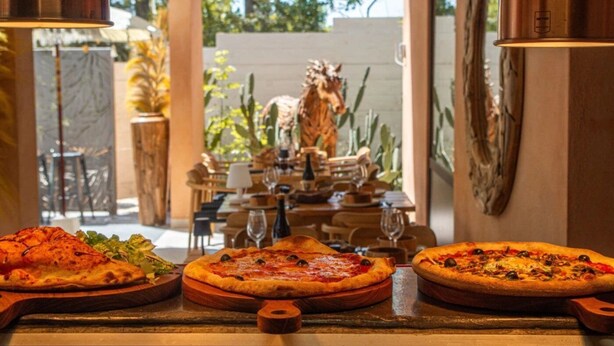 A restaurant with three pizzas on wooden boards, a dining table with chairs, and a statue in the background. - ZEcamping 3* piscine pet friendly mobil-home 33m2 6 pers (Tuchan)