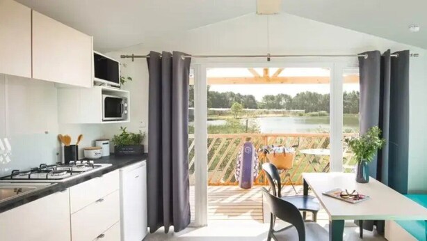 A modern kitchen with a gas stove, microwave, and cabinets. There is a dining area with a table and chairs, and a view of a lake through large windows. - ZEcamping 3* piscine pet friendly mobil-home BBQ 5 pers (Sandrans)