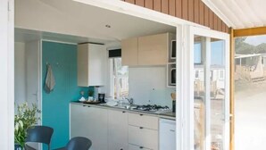 A compact kitchen with white cabinets, a sink, and a stove. There is a dining area with two chairs and a table. The room has a view of a residential area through a glass door. - ZEcamping 3* piscine pet friendly mobil-home BBQ 5 pers (Sandrans)