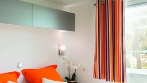 A modern bedroom with a bed, pillows, a bedside table with a lamp, a window with striped curtains, and a view of the outdoors. - ZEcamping 3 étoiles piscine chiens acceptés mobil-home BBQ 30m2 6 pers (Sandrans)