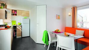 A compact kitchen with white cabinets, a refrigerator, and a dining area with a table and chairs. - ZEcamping 3 étoiles piscine chiens acceptés mobil-home BBQ 30m2 6 pers (Sandrans)