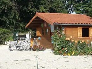 A wooden building with a tiled roof, bicycles parked in front, and a flower bed with various flowers. - ZEcamping 3* piscine animaux acceptés mobil-home BBQ 8 pers (Sandrans)