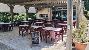 Un café en plein air avec des tables et des chaises en bois, un espace de repos couvert et un menu écrit à la craie sur un tableau noir.