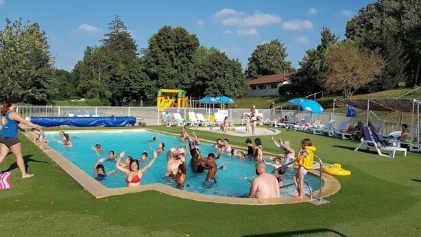Piscina con gente disfrutando del agua, rodeada de césped y árboles.