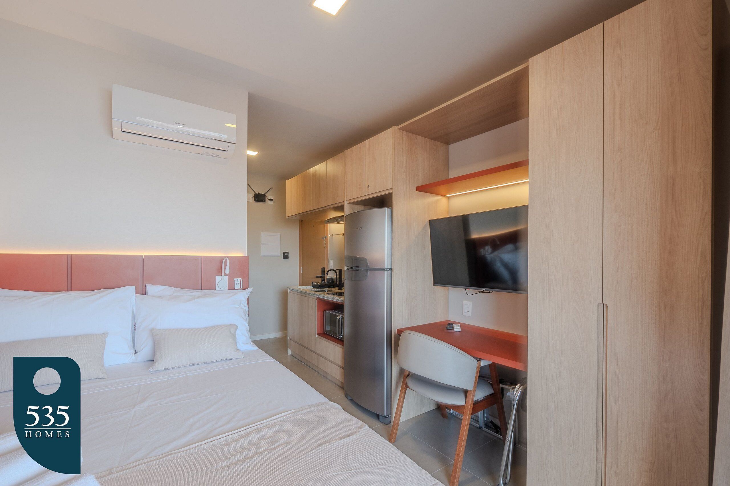A hotel room with a bed, a desk, a chair, a TV, a refrigerator, and a kitchen area.