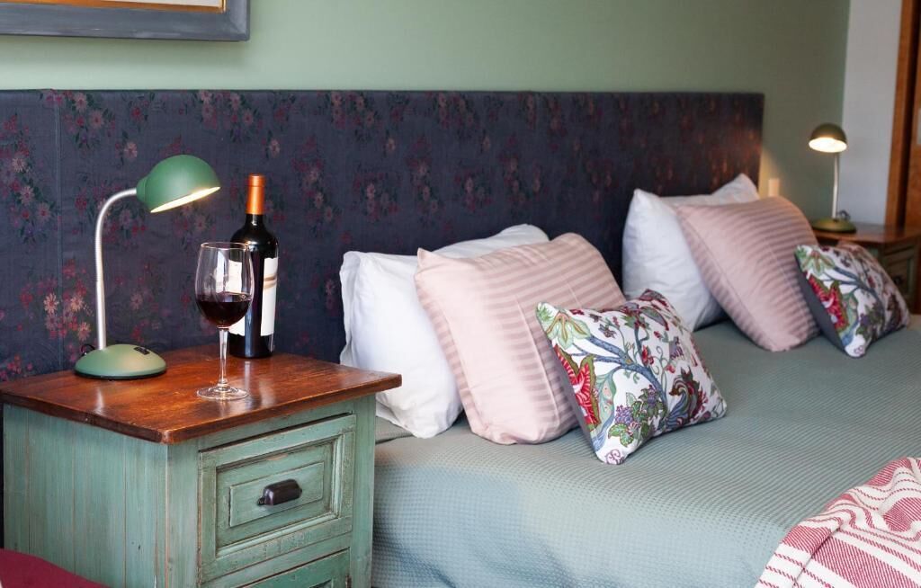 A bedroom with a floral-patterned headboard, a wooden nightstand with a lamp, a bottle of wine, and a glass. There are multiple pillows on the bed.