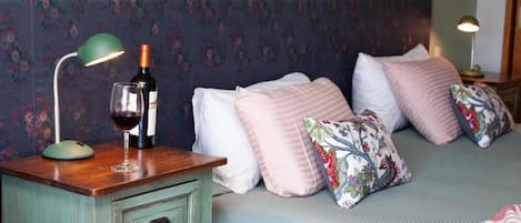 A bedroom with a floral-patterned headboard, a wooden nightstand with a lamp, a bottle of wine, and a glass. There are multiple pillows on the bed.