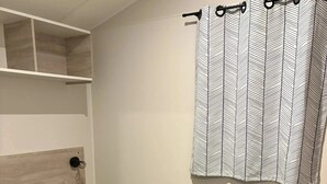 Una cama individual con colchón gris y almohada negra, cabecera de madera, mesita de noche con lámpara y cortina estampada.