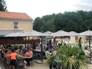 An outdoor seating area with tables and umbrellas, surrounded by trees and a building with a tiled roof. - Camping 3 étoiles - Piscine  - ccbfa0h (Saint-Mandé-sur-Brédoire)