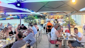 Uma área de refeições ao ar livre com mesas e guarda-sóis, um palco com músicos e vista para o mar.