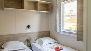 A bedroom with two beds, wooden headboards, a window with blinds, and a small shelf above the beds. - ZEcamping 3 étoiles pataugeoire piscine locatif TV LV clim 33m2 6 pers (Saint-Remy-de-Provence)
