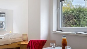 Un dormitorio con cama, mesita de noche, lámpara, ventana con vista a árboles y una mesa con manzanas y una botella.