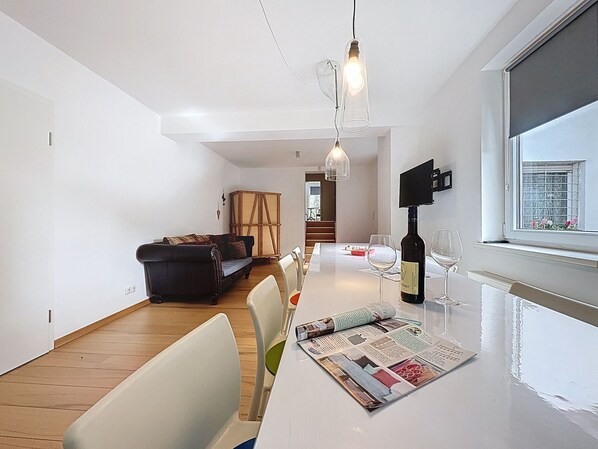 A modern living room with a dining area, a sofa, a table with a magazine, and a wine bottle.