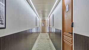 Ein langer, schmaler Korridor mit Teppichboden, gesäumt von Wänden mit gerahmten Bildern und einer Holztür auf der rechten Seite.