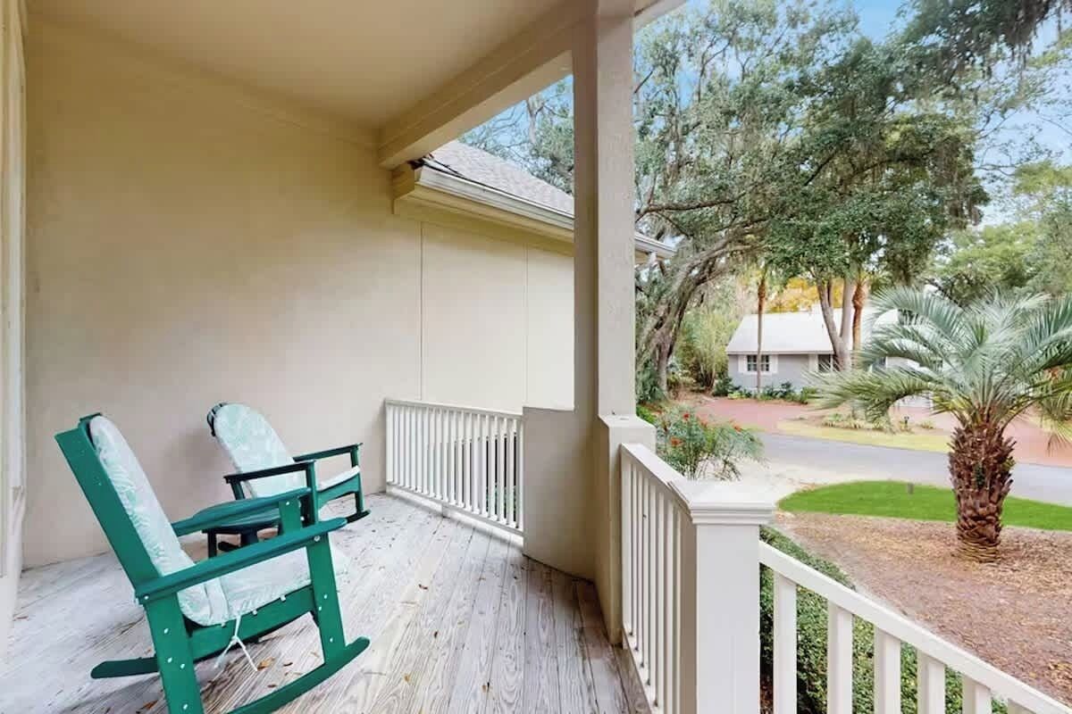 A porch with two rocking chairs, a white railing, and a view of a residential street with trees and a palm tree.