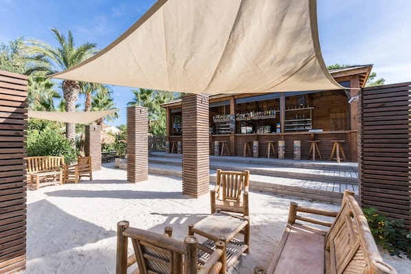 An outdoor seating area with wooden furniture and a shaded canopy, a bar with wooden shelves, and palm trees in the background.