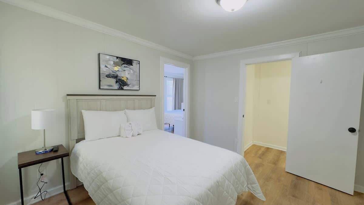 A bedroom with a bed, a bedside table with a lamp, a painting on the wall, and a door leading to another room.