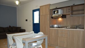 A compact kitchen with a microwave, stove, and sink. There is a dining table with chairs and a blue door in the background. - Residence Villaggio Petruso (Balestrate)