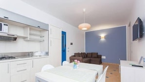 A compact kitchen with white cabinets, a dining table with chairs, a blue door, and a brown sofa in a living area with a light fixture. - Residence Villaggio Petruso (Balestrate)