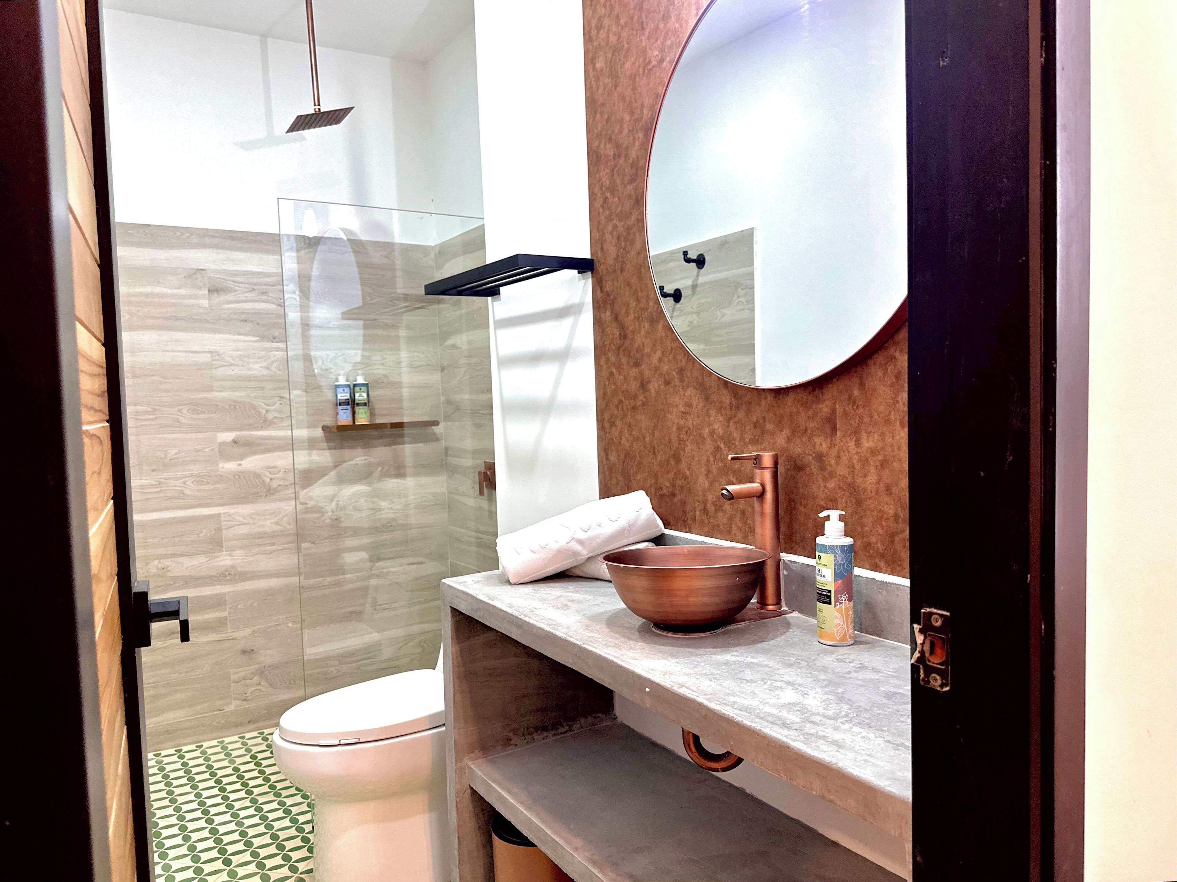 A modern bathroom with a glass shower enclosure, a round mirror, a copper sink, and a toilet.