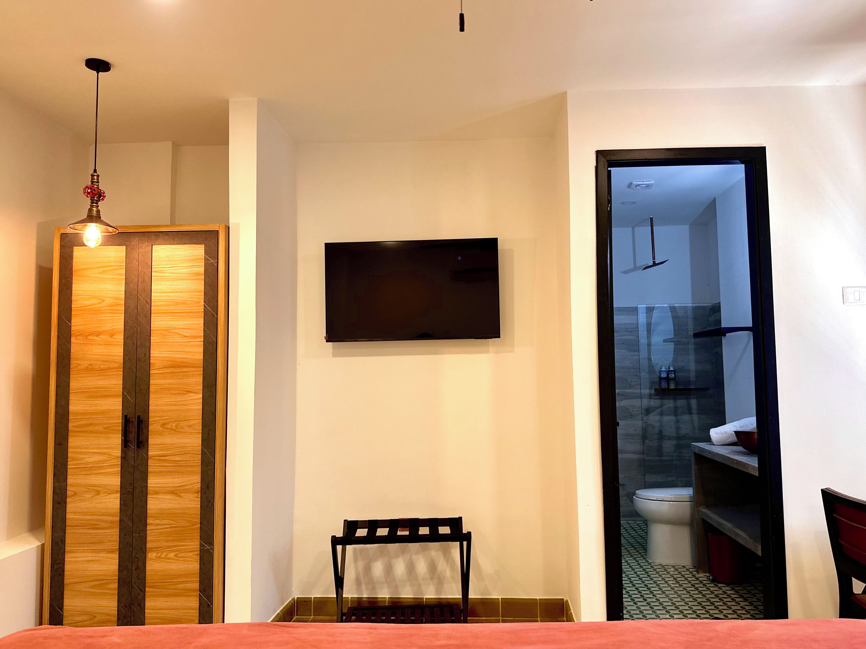 A hotel room with a wooden wardrobe, a flat-screen TV mounted on the wall, a chair, and a view of the bathroom through an open door.