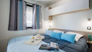 A bedroom with a bed, pillows, a hat, and a magazine. - ZEcamping 4* parc aquatique animations locatif TV LV clim 4 pers (Sérignan)