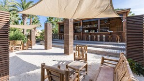 An outdoor seating area with wooden furniture and a shaded canopy, a bar with wooden shelves, and palm trees in the background. - ZEcamping 4* parc aquatique animations locatif TV LV clim 4 pers (Sérignan)
