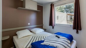 Ein Einzelbett mit weißer und blauer Bettwäsche, einem gestreiften Kopfkissen und einer Fernbedienung darauf. Ein Fenster mit braunen Vorhängen und Blick ins Freie.