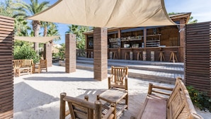 Un área de estar al aire libre con muebles de madera y un toldo sombreado, un bar con estantes de madera y palmeras al fondo.