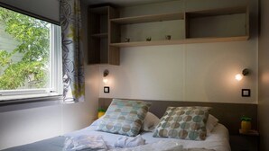 Una cama bien hecha con almohadas, una ventana con cortinas y estantes en la pared con objetos decorativos.