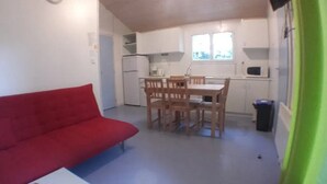 Ett kompakt vardagsrum med en röd soffa, ett matbord med stolar, en köksdel med kylskåp och mikrovågsugn samt ett fönster med gardiner.