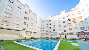 A swimming pool surrounded by green artificial grass, with a multi-story building in the background. - HomeHolidaysRentals Olimpia - Costa Barcelona (Pineda de Mar)