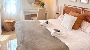 Una habitación de hotel con cama, escritorio, silla, espejo redondo, lámpara de pared y taburete de mimbre.