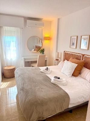 A hotel room with a bed, a desk, a chair, a round mirror, a wall-mounted lamp, and a wicker stool. - BONANZA 1 SUNNY BEDROOM APARTMENT, NEW 2026! (Arroyo de la Miel)
