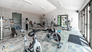 Moderni kuntosali, jossa on monipuolinen valikoima kuntoiluvälineitä, kuten kuntopyöriä ja painonnostolaitteita, sekä suuri ikkuna, josta tulee runsaasti luonnonvaloa.